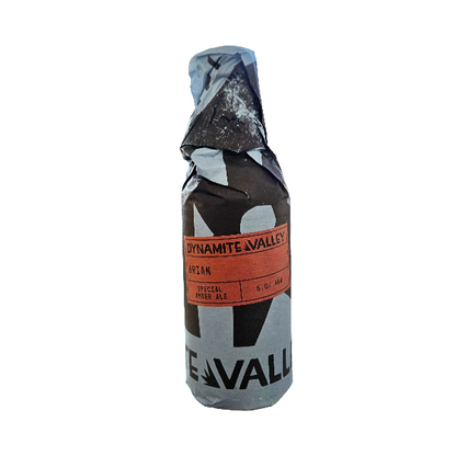 A bottle of Dynamite Valley Ale wrapped in a camouflage-patterned paper sleeve, isolated on a white background.