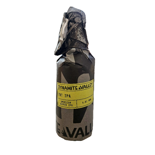 A single packaged beer standing upright on a plain white background. The package is wrapped in dark gray and black camouflage paper with a wide yellow label reading 'DYNAMITE VALLEY' and smaller text 'TNT SPA' and 'AMERICAN TOPPED SPA'.