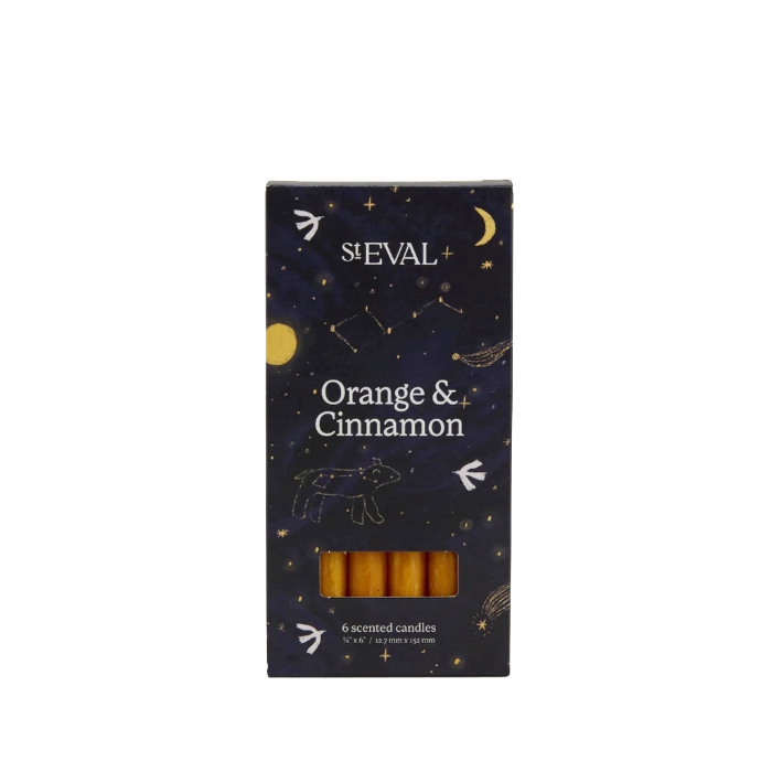 A boxed set of six St Eval Orange & Cinnamon scented candles. The packaging is a dark navy blue with a celestial pattern of stars, moons, and constellations, and features a clear window at the bottom showing the golden-yellow wax candles inside.