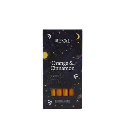 A boxed set of six St Eval Orange & Cinnamon scented candles. The packaging is a dark navy blue with a celestial pattern of stars, moons, and constellations, and features a clear window at the bottom showing the golden-yellow wax candles inside.