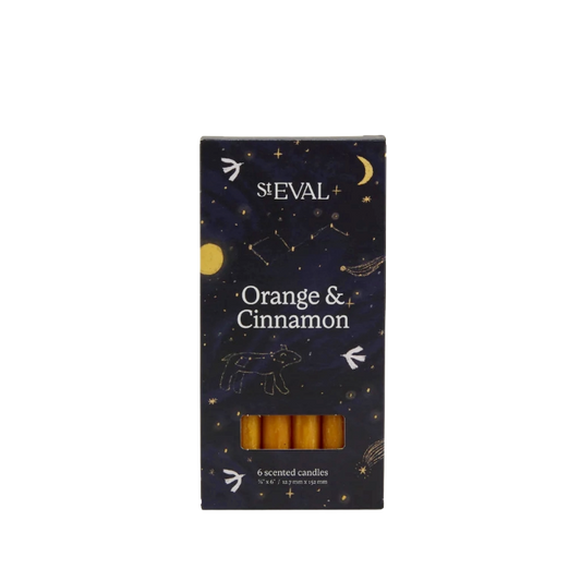 A boxed set of six St Eval Orange & Cinnamon scented candles. The packaging is a dark navy blue with a celestial pattern of stars, moons, and constellations, and features a clear window at the bottom showing the golden-yellow wax candles inside.