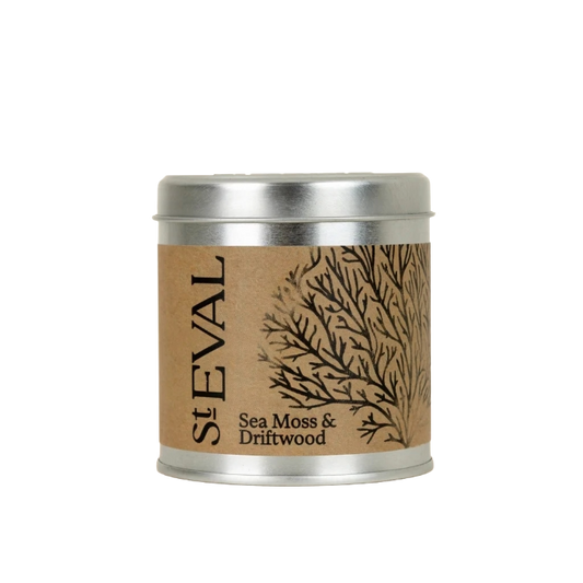 A St Eval Sea Moss & Driftwood scented candle in a round silver metal tin with a brown kraft paper label featuring a black branch illustration.